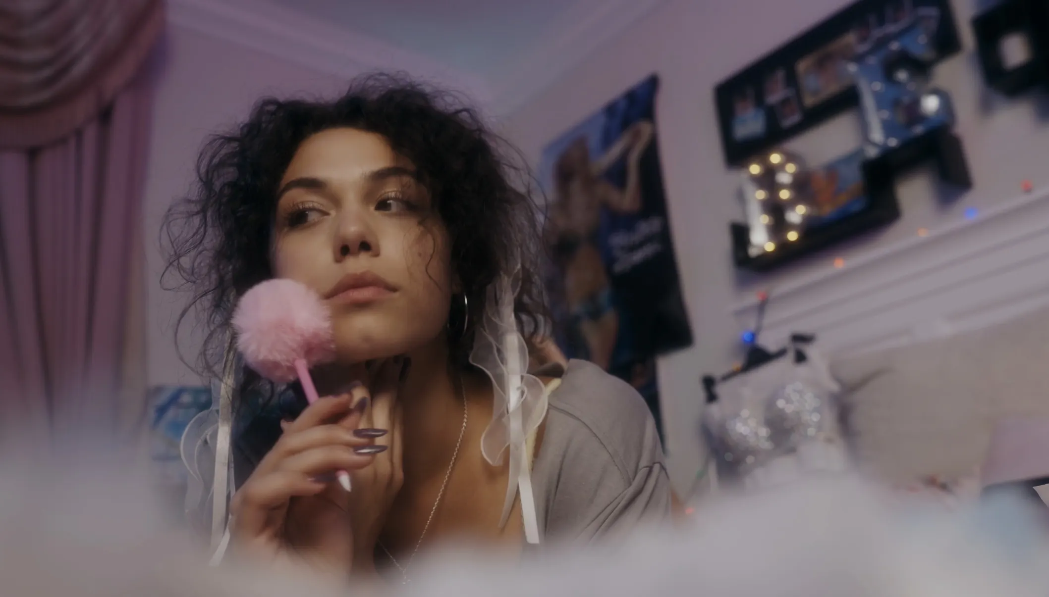 Film still from the Play Pretend music video: Bella Valdes in soft pink light holding a pom-pom pen, gaze off camera, in a teenage bedroom set.